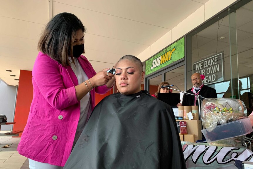 A woman wearing a face mask shaves the head of a girl.