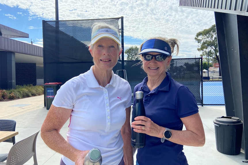 Two woman standing smiling at the camera wearing tennis sport clothing, including visors. 