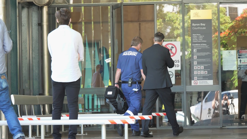 A man in a blue 'paramedic' uniform carrying a bag into a building. Two other men standing nearby.