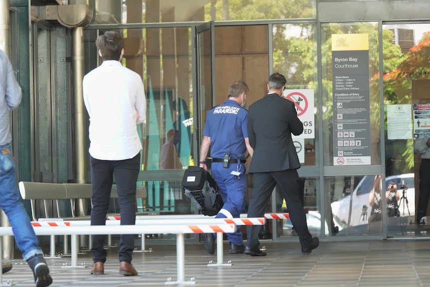 A man in a blue 'paramedic' uniform carrying a bag into a building. Two other men standing nearby.