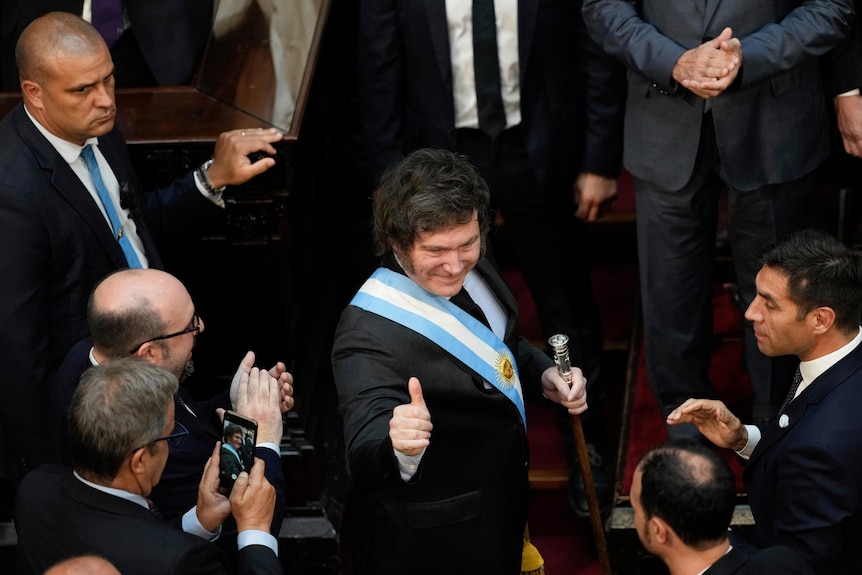 A middle-aged man with thick dark hair, wearing a blue and white sash, gives a thumbs-up in a crowd of suit-wearing men.