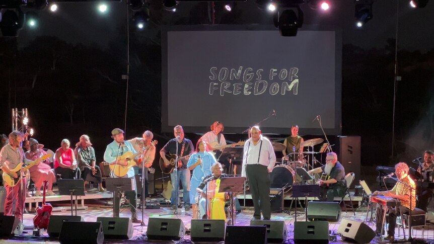 Singers and a live band perform on stage with 'Songs for Freedom' projected behind them.