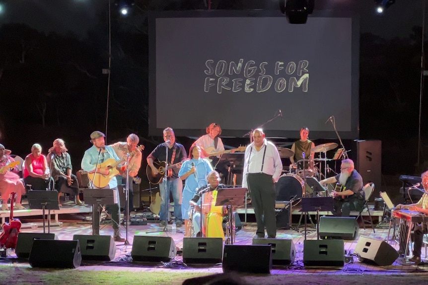 Singers and a live band perform on stage with 'Songs for Freedom' projected behind them.