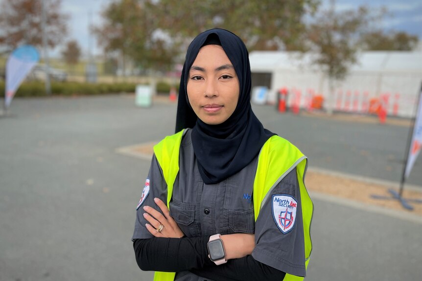A young woman wearing a headscarf and a hi-vis vest stands outside with her arms crossed.