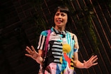 The actor stands on stage, in a brightly printed dress, in front of a moody, leafed backdrop.