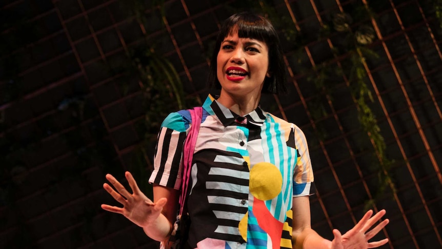 The actor stands on stage, in a brightly printed dress, in front of a moody, leafed backdrop.