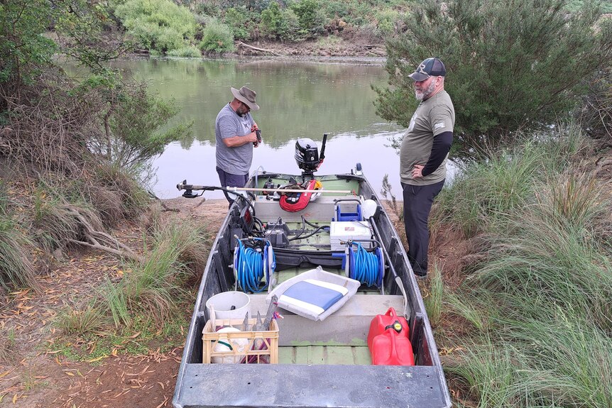 Two men stand next to a boat on a riverbank. The boat is loaded with equipment.
