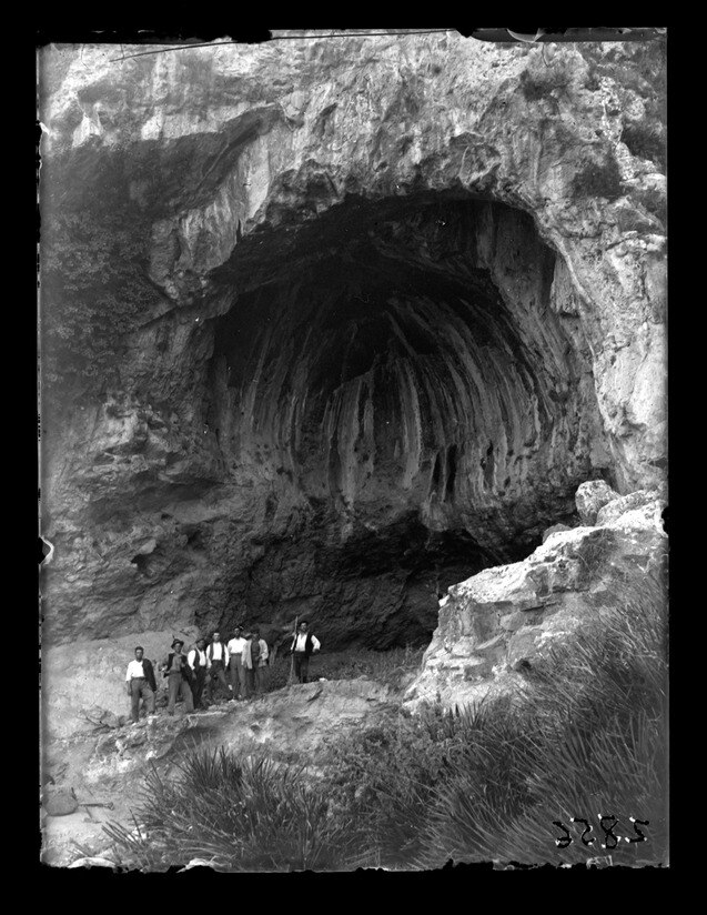 A black and white photo of a group of men standing in front of a cave.