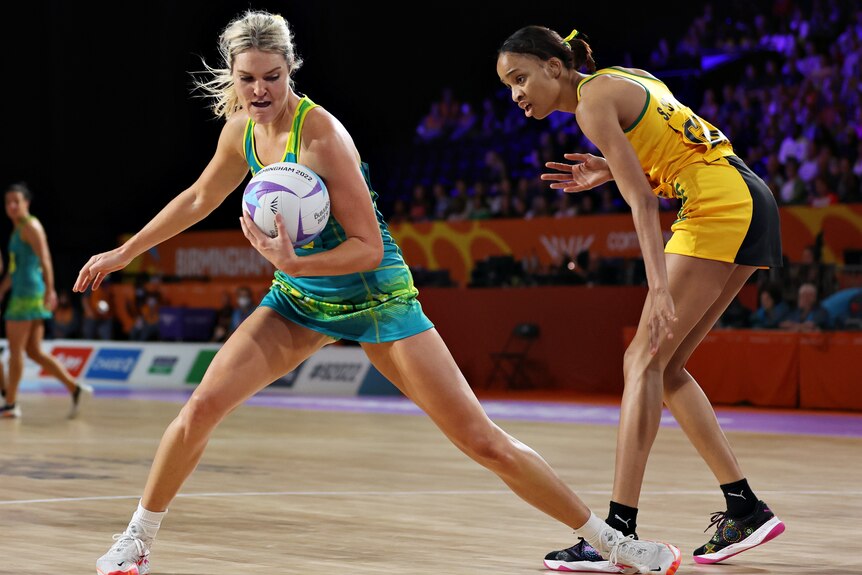 An Australian netball player holds the ball with her left hand while standing next to a Jamaican opponent.