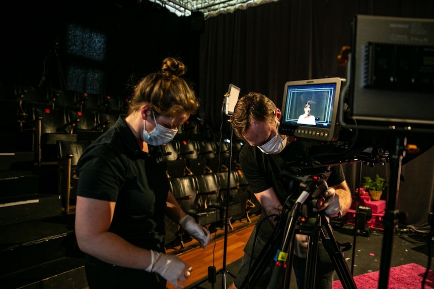 Two people wearing masks set up filming equipment in a darkened theatre.