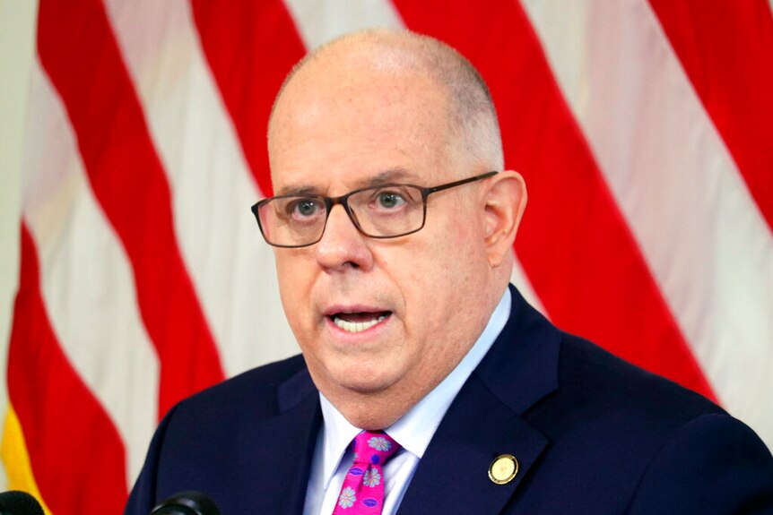 A bad man with glasses, a blue suit jacket and a pink floral tie stands in front of a US flag.