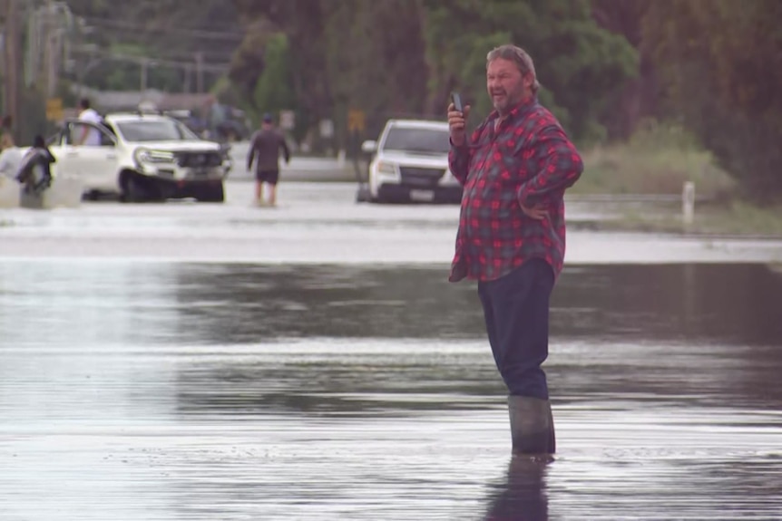 A man in a red shift talks on the phone while standing in a flooded road.