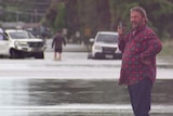 A man in a red shift talks on the phone while standing in a flooded road.