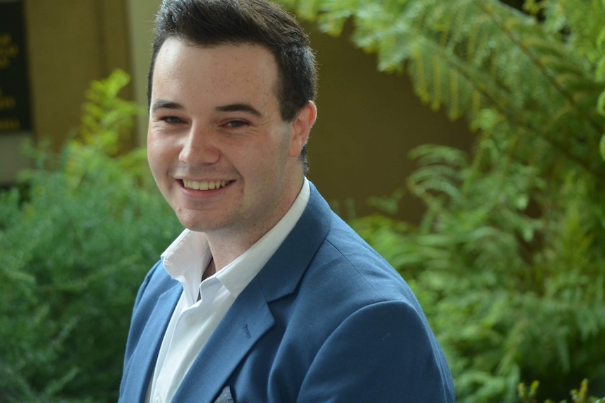 Ben Dudman smiles at the camera while wearing a suit. He's standing in front of some ferns.