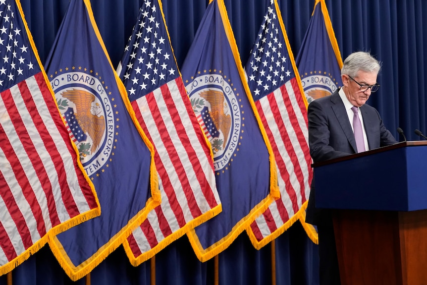 James Powell stands on a podium in front of a blue curtain and a row of American flags. 