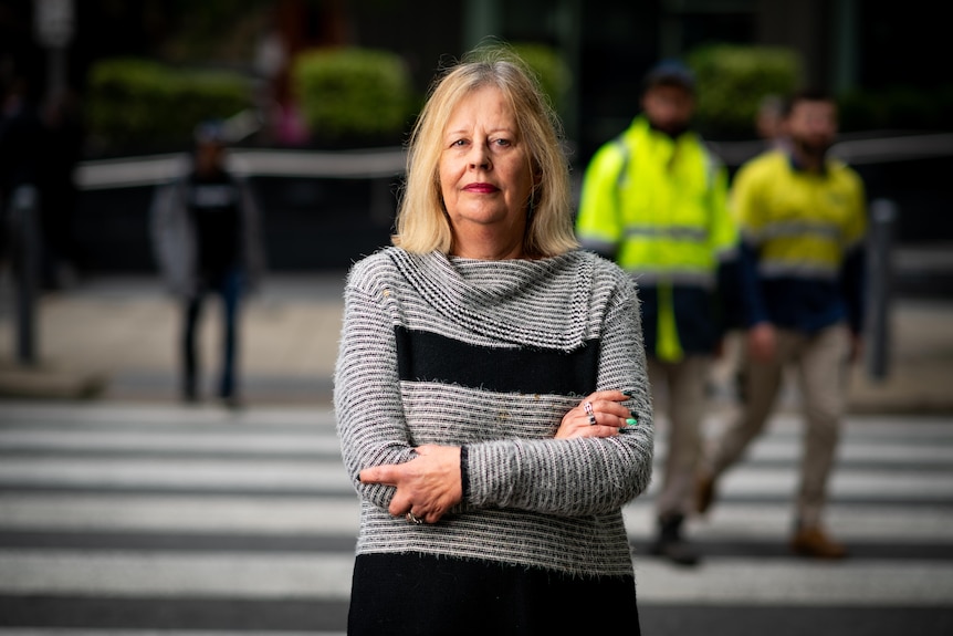 A woman stands with a serious expression and her arms folded with people walking across a street crossing behind her