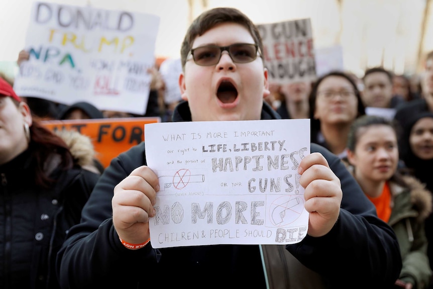 A boy holds a sign reading "what is more important our right to life, liberty... or more guns?"