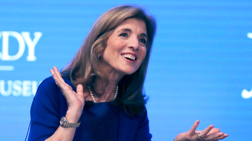 Caroline Kennedy smiling and gesturing against a blue background.