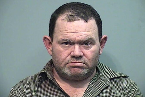 An unshaven, thickset man in a buttoned shirt stares at the camera for a mugshot.