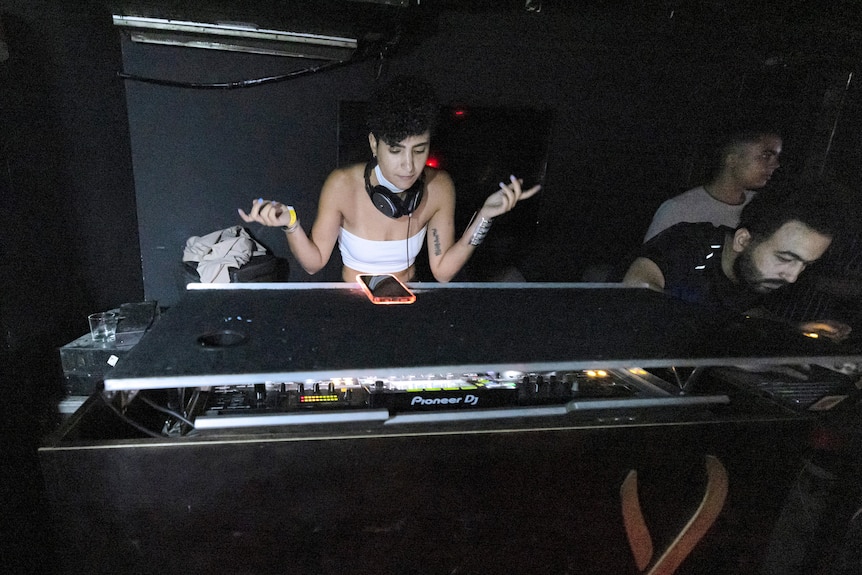 a woman with headphones around her neck raises her hands up to her side as she stands at a DJ console next to two men