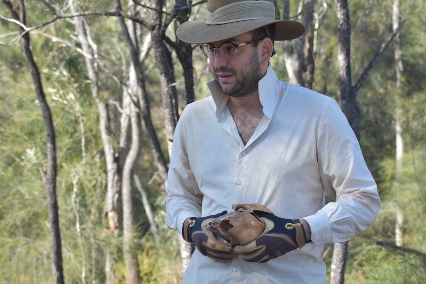 Julie wearing a white button up shirt and hat holding a specimen, trees behind.
