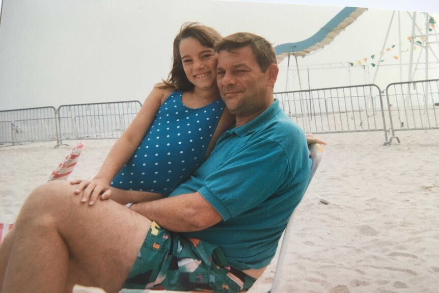 A girl in a blue and white polkadot swimsuit hugs a man in a blue shirt as they sit on a chair on the beach.