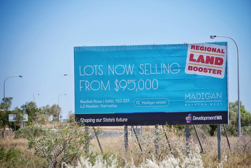 A large blue 'Lots Now Selling' billboard by the side of the road in front of vacant land.