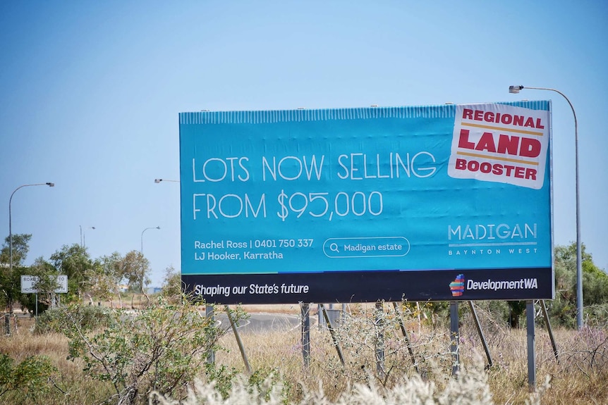 A large blue 'Lots Now Selling' billboard by the side of the road in front of vacant land.