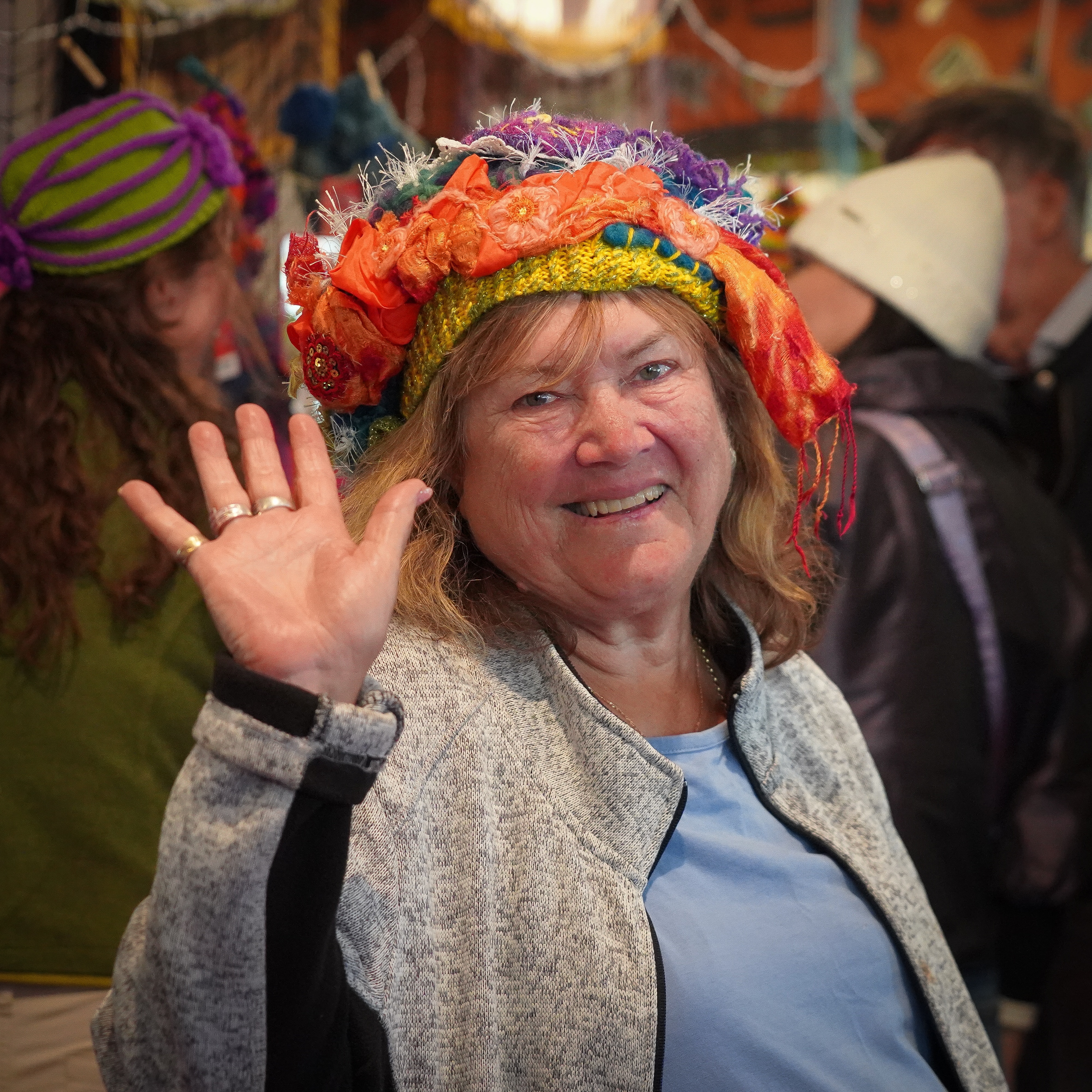 A woman at waving and smiling at the camera while wearing a rainbow-coloured beanie.