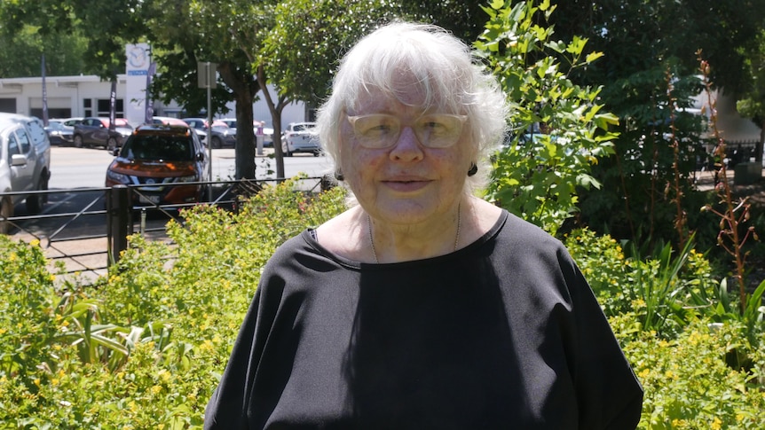An elderly woman with grey hair in a black top and clear glasses standing in front of green bus, slight smile.