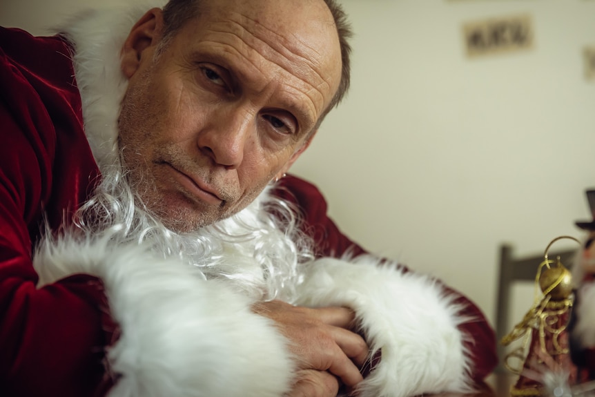 An inebriated looking man in a santa suit leans on a counter.