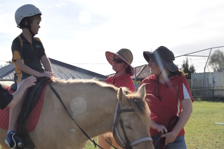 Two volunteers wearing red assist a young boy in a green and yellow shirt who sits on top a beige horse.