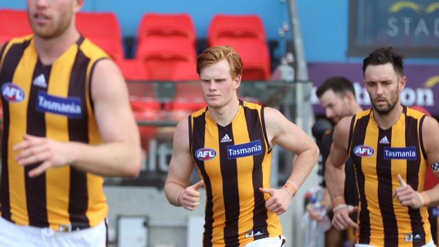 A young man with red hair runs next to teammates wearing a brown and gold uniform. 