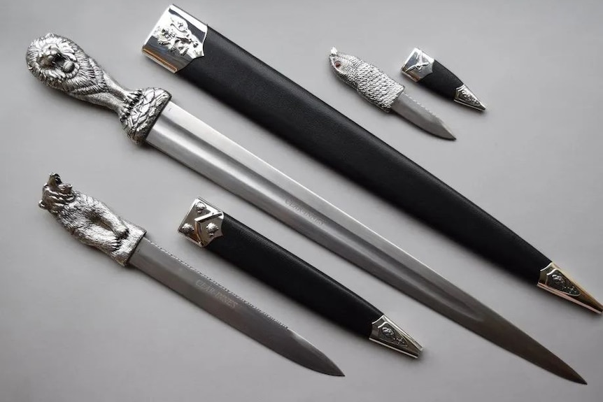 Three silver daggers of various length with lion heads on the handles and black sheaths on a table.