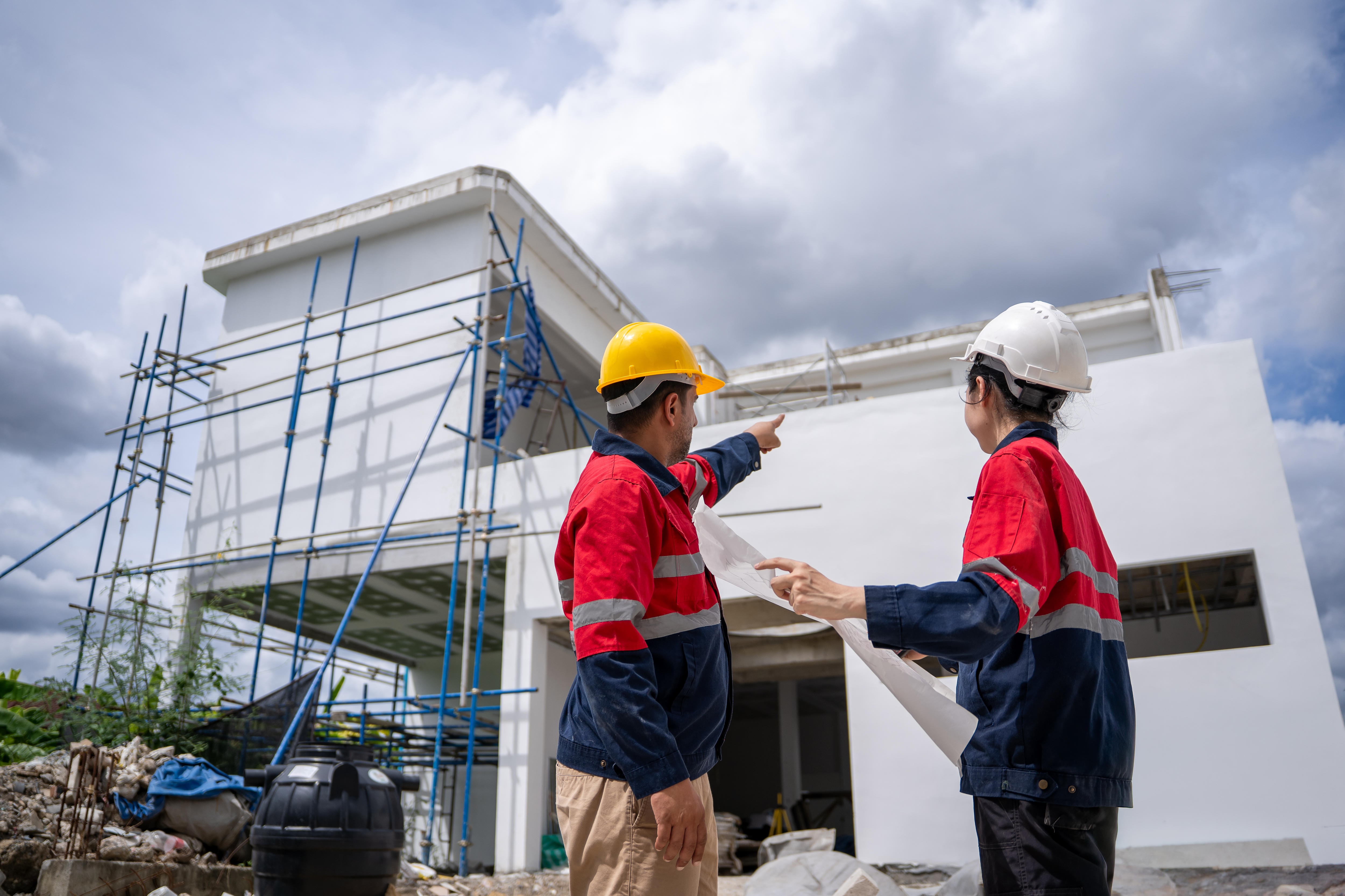 Two builders are inspecting a construction site. They both wear hard hats. A male is pointing, the female holding plans.