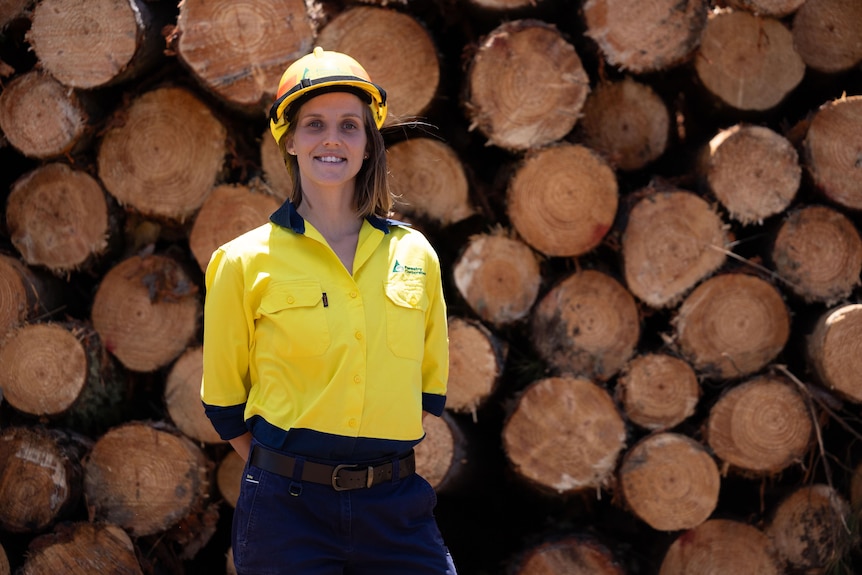 A woman in high vis stands in front of a pile of timber logs 
