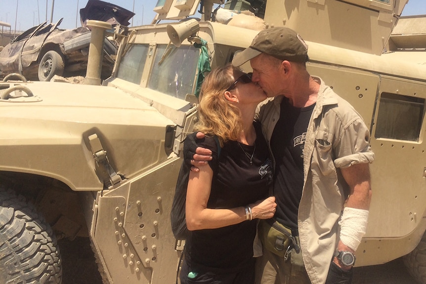 A couple, the man with a bandaged arm, kisses in front of a dusty armoured vehicle.