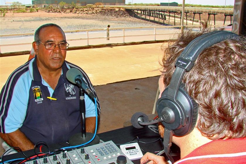 Man wearing blue shirt sits inside a studio, speaking to another man with headphones on