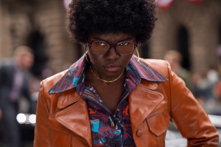 A Black woman with an afro, wears an orange leather jacket, a purple patterned shirt and glasses & looks staunchly at the camera