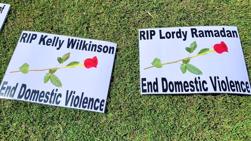 Two signs lying on grass saying RIP Kelly Wilkinson and RIP Lordy Ramadan, end domestic violence