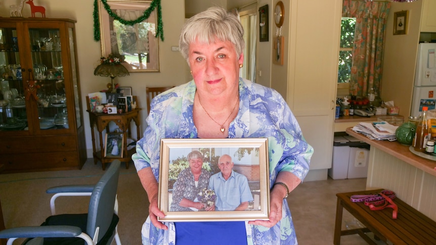 Woman with short grey hair wearing blue and white top holding a photo of herself and her husband