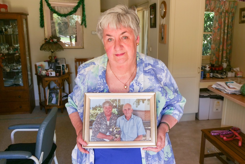 Woman with short grey hair wearing blue and white top holding a photo of herself and her husband
