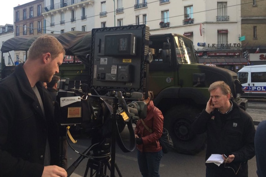 Williams standing on street with hand to ear looking to camera with army and police trucks in background.