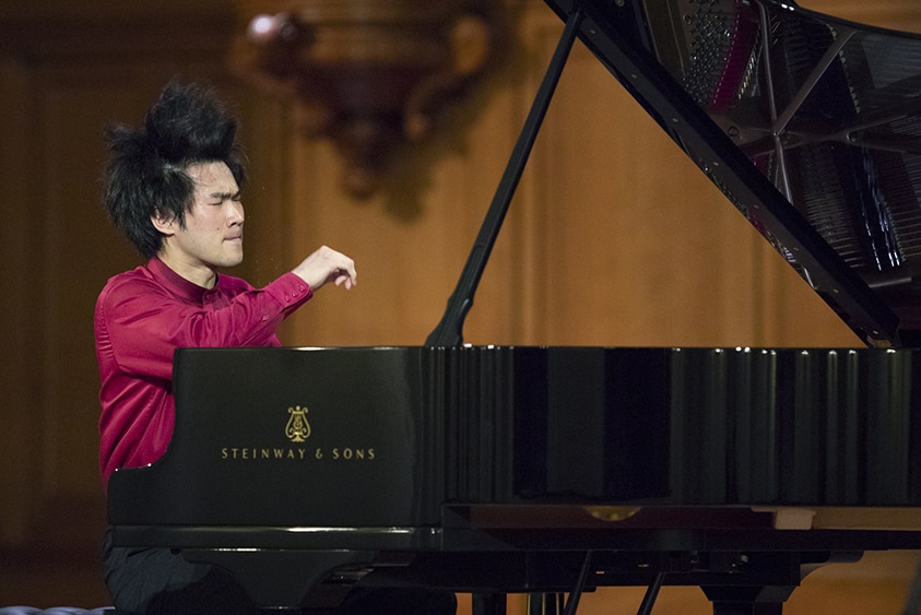 A pianist performs on a grand piano. One arm is dramatically raised and his hair is leaping upwards.