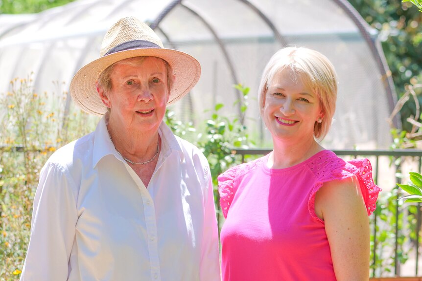 An older woman wearing a straw hat smiles next to a smiling younger woman in a bright pink top. They're standing in a garden.