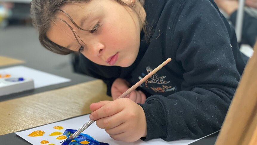 A girl holding a paintbrush looks down at a piece of paper where she is painting with blue and yellow