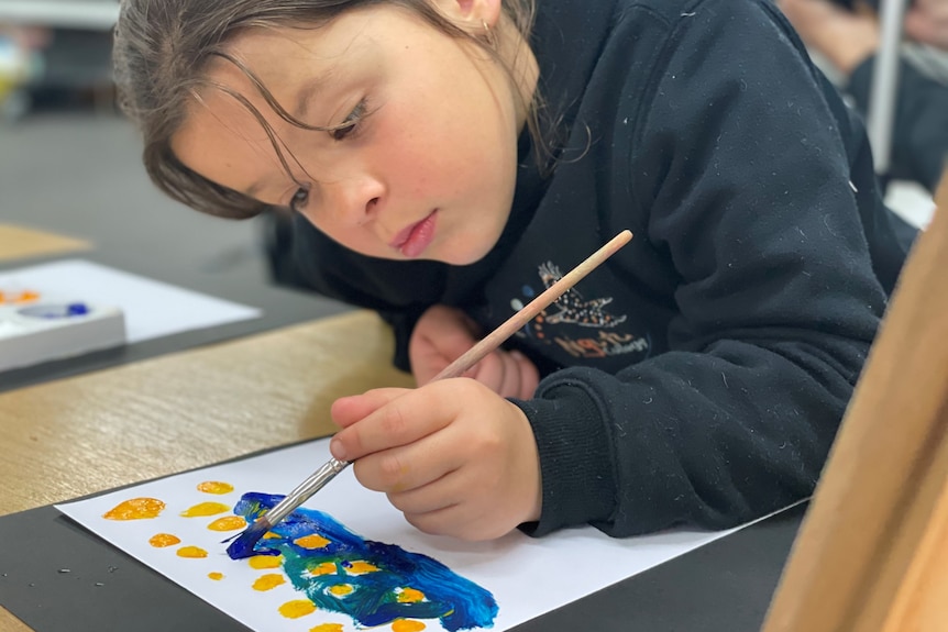 A girl holding a paintbrush looks down at a piece of paper where she is painting with blue and yellow