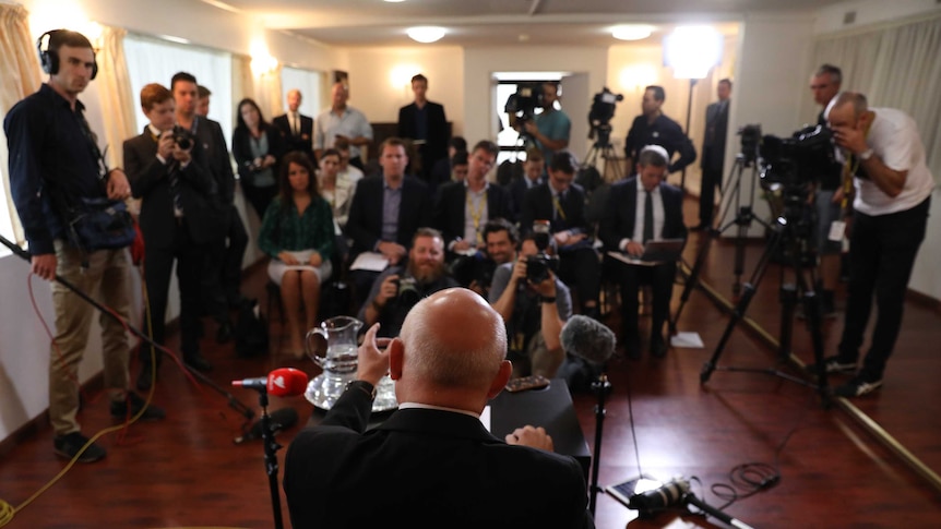 Grigory Logvinov, as seen from behind, gestures to a watching crowd of journalists. There are several cameras in the room.