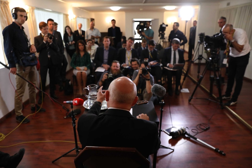 Grigory Logvinov, as seen from behind, gestures to a watching crowd of journalists. There are several cameras in the room.
