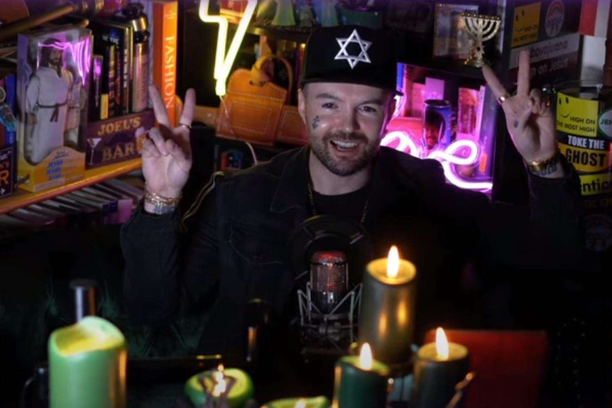 Man wearing black jacket and black cap holds up two finger peace sign with both hands in dark neon lit room
